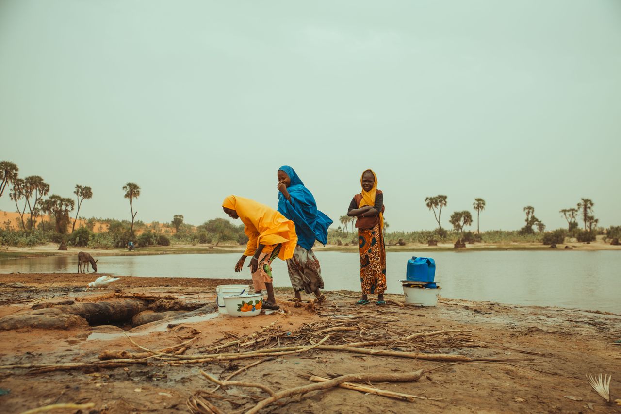 Three women in colorful attire gather water by an African riverbank, depicted in a natural setting.
