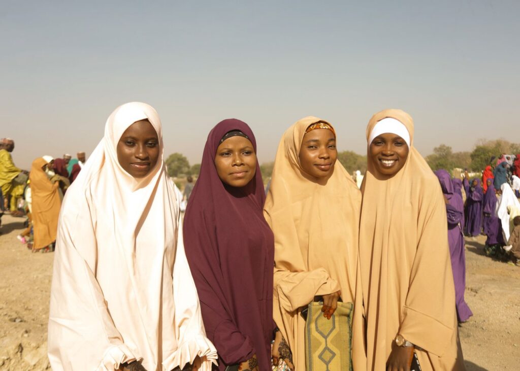 Four women wearing traditional hijabs smiling outdoors on a sunny day, creating a warm and joyful atmosphere.